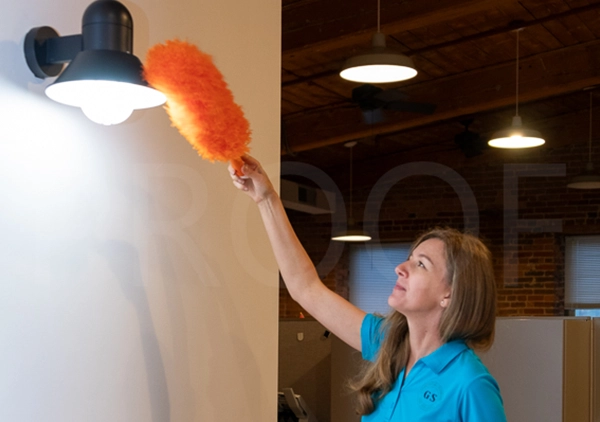A woman is cleaning a light fixture with a mop, standing on a ladder in a well-lit space